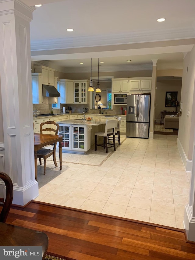 kitchen featuring light wood finished floors, a kitchen bar, backsplash, appliances with stainless steel finishes, and decorative columns