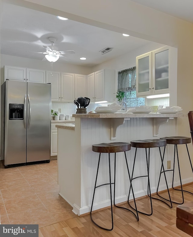 kitchen with a kitchen breakfast bar, ceiling fan, stainless steel fridge with ice dispenser, glass insert cabinets, and white cabinetry