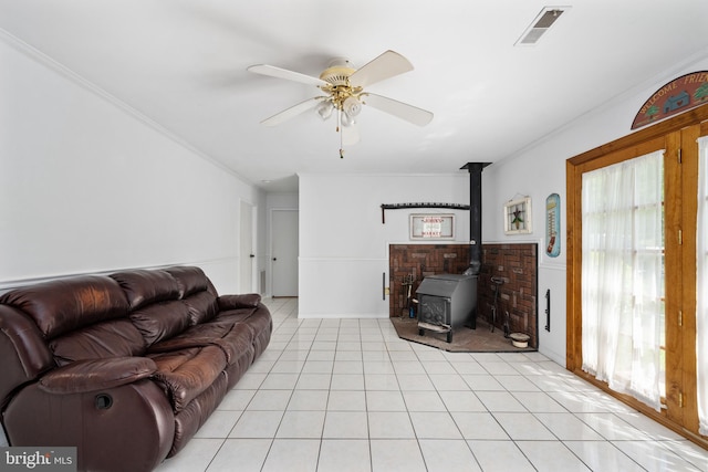 living area featuring light tile patterned flooring, ceiling fan, a wood stove, visible vents, and ornamental molding