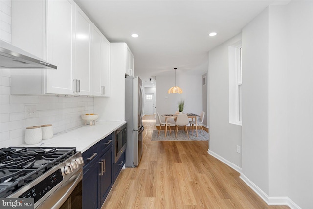 kitchen with wall chimney range hood, blue cabinets, appliances with stainless steel finishes, white cabinetry, and light wood-style flooring