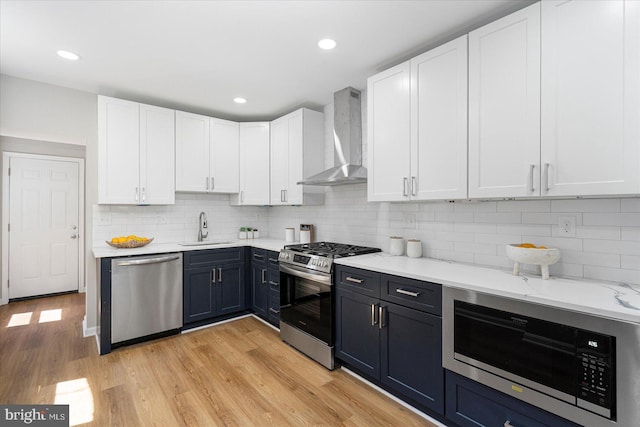 kitchen with wall chimney range hood, appliances with stainless steel finishes, a sink, light wood-style flooring, and white cabinetry
