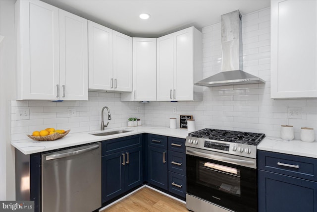 kitchen featuring wall chimney range hood, a sink, blue cabinetry, stainless steel appliances, and white cabinetry
