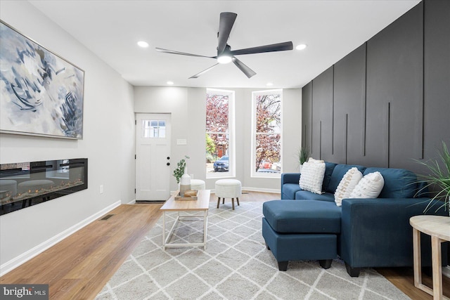 living room with a glass covered fireplace, light wood-style flooring, a ceiling fan, baseboards, and recessed lighting