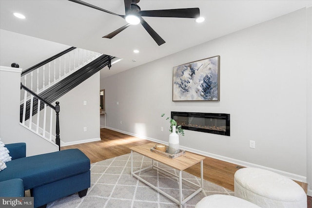 living area featuring a glass covered fireplace, stairs, a ceiling fan, baseboards, and wood finished floors