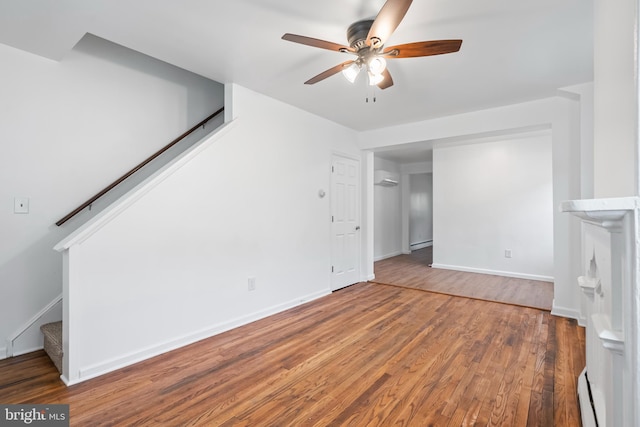 unfurnished living room with stairway, a baseboard radiator, wood finished floors, ceiling fan, and baseboards