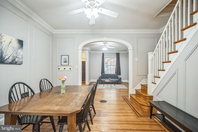 dining area featuring arched walkways, ceiling fan, crown molding, light wood-type flooring, and stairs