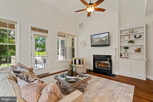 living area with ceiling fan, visible vents, dark wood-style floors, a glass covered fireplace, and a high ceiling