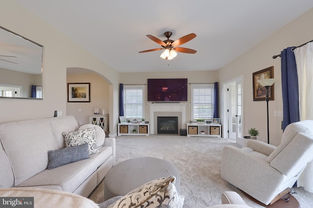 carpeted living area featuring a fireplace with flush hearth, arched walkways, and ceiling fan