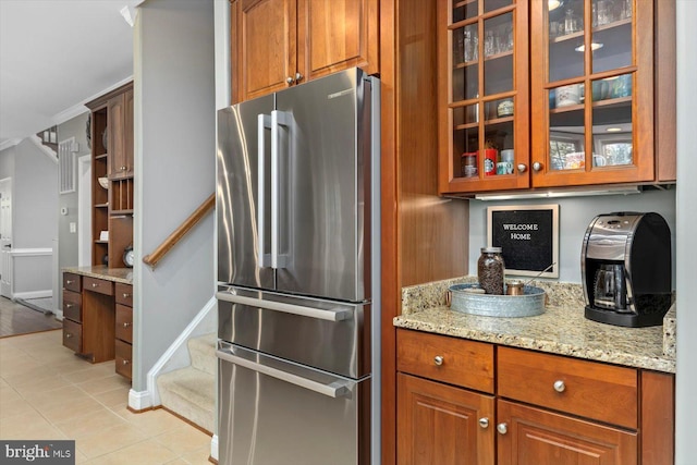 kitchen with brown cabinetry, light stone countertops, freestanding refrigerator, glass insert cabinets, and light tile patterned flooring