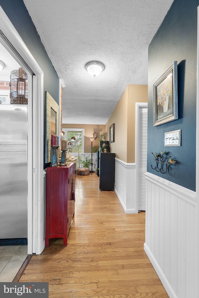 hall featuring light wood-style flooring, a textured ceiling, and a wainscoted wall