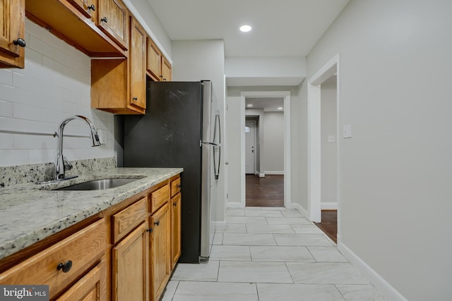 kitchen featuring a sink, freestanding refrigerator, light stone countertops, brown cabinetry, and tasteful backsplash
