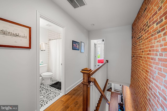 hall with an accent wall, an upstairs landing, brick wall, wood-type flooring, and visible vents