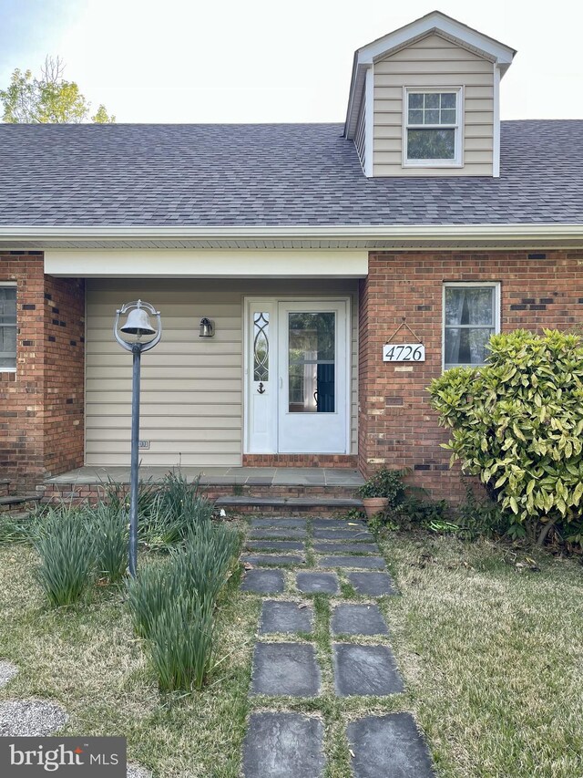 doorway to property with covered porch, brick siding, and a shingled roof