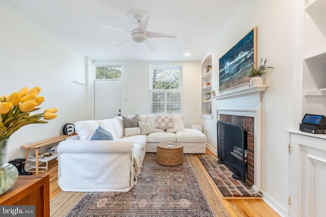living area featuring a brick fireplace, a ceiling fan, built in features, and wood finished floors