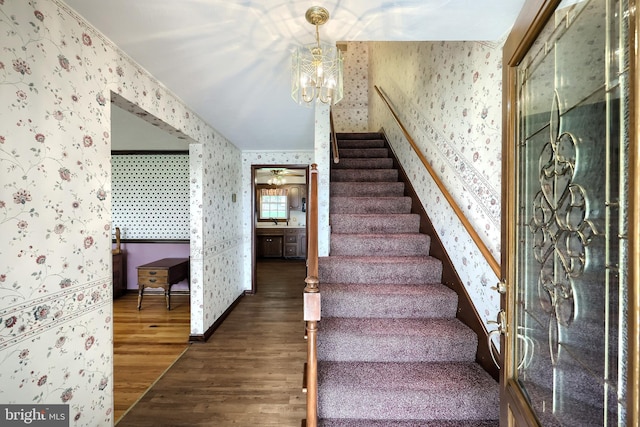 stairway featuring baseboards, a notable chandelier, wood finished floors, and wallpapered walls