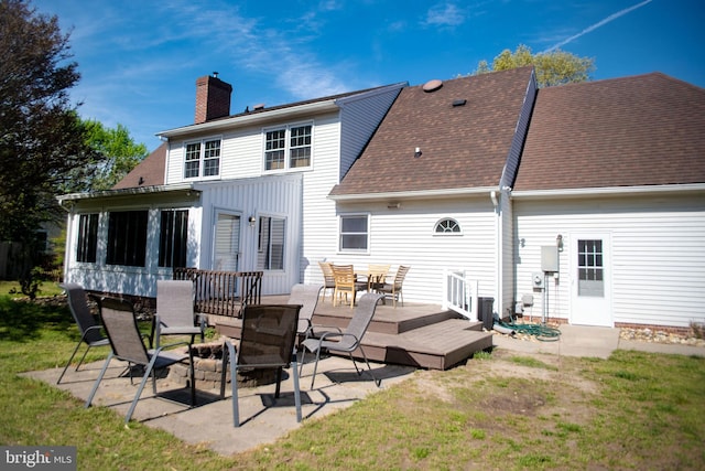 back of property with a deck, roof with shingles, a chimney, a lawn, and outdoor dining space