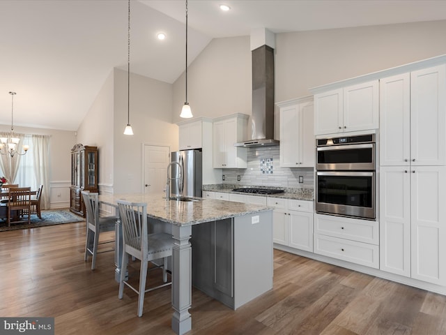 kitchen featuring an island with sink, stainless steel appliances, wall chimney range hood, a sink, and high vaulted ceiling