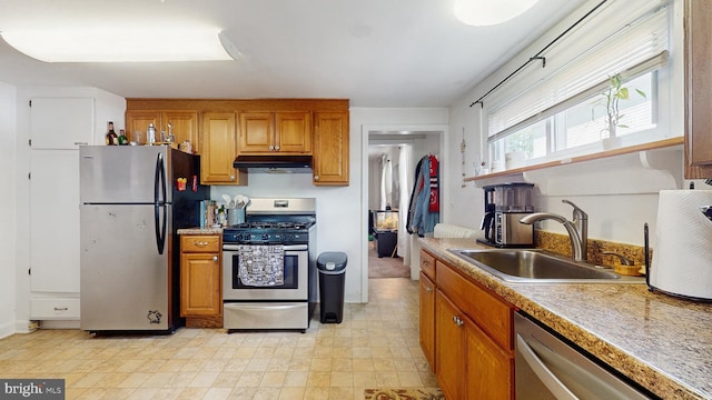 kitchen with stainless steel appliances, under cabinet range hood, a sink, brown cabinetry, and light countertops