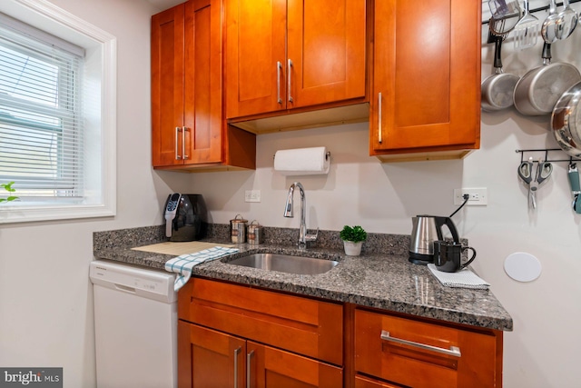 kitchen featuring white dishwasher, a sink, brown cabinets, and dark stone counters