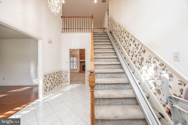 stairway with tile patterned floors, wallpapered walls, wainscoting, a towering ceiling, and a brick fireplace