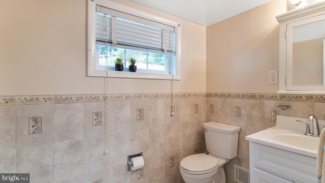 half bath featuring visible vents, tile walls, wainscoting, vanity, and toilet
