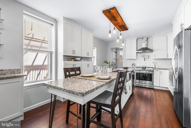 kitchen featuring stainless steel appliances, backsplash, light stone counters, wall chimney range hood, and dark wood-type flooring