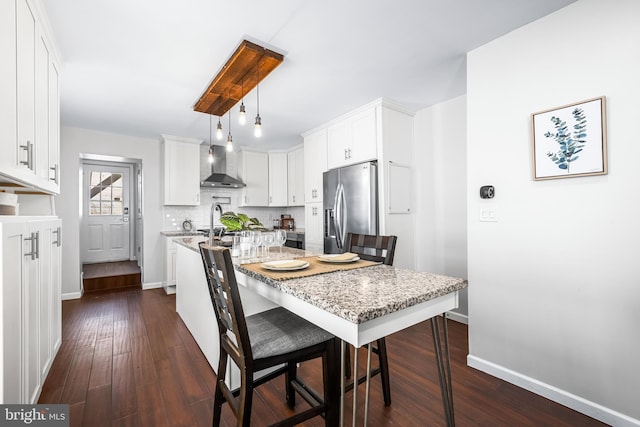 kitchen with wall chimney range hood, stainless steel fridge with ice dispenser, white cabinetry, light stone countertops, and dark wood-type flooring