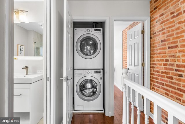 washroom with dark wood-style floors, a sink, brick wall, stacked washer and dryer, and laundry area