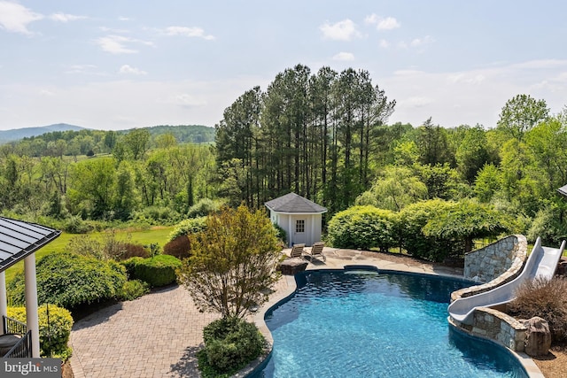 outdoor pool featuring a water slide, a view of trees, a storage structure, an outdoor structure, and a patio area