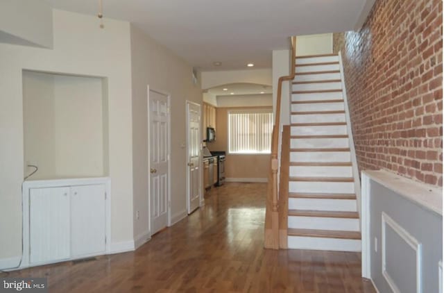 hall featuring wood finished floors, arched walkways, baseboards, stairs, and brick wall
