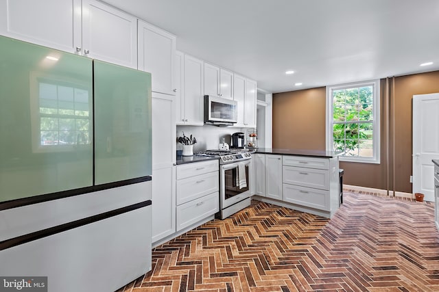 kitchen featuring stainless steel gas range, white cabinets, dark countertops, brick floor, and refrigerator with glass door