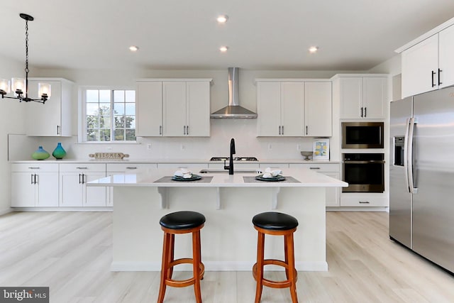 kitchen featuring light wood finished floors, a kitchen island with sink, wall chimney range hood, light countertops, and appliances with stainless steel finishes