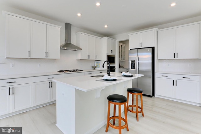 kitchen featuring light wood finished floors, a sink, wall chimney range hood, a breakfast bar, and appliances with stainless steel finishes