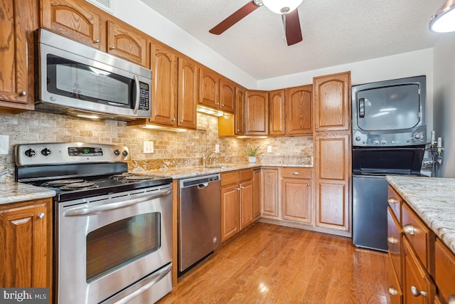 kitchen featuring a ceiling fan, stacked washer / dryer, light wood-type flooring, appliances with stainless steel finishes, and light stone countertops