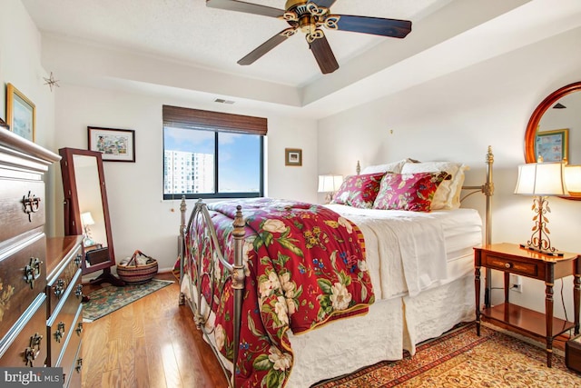 bedroom featuring a raised ceiling, wood finished floors, visible vents, and a ceiling fan