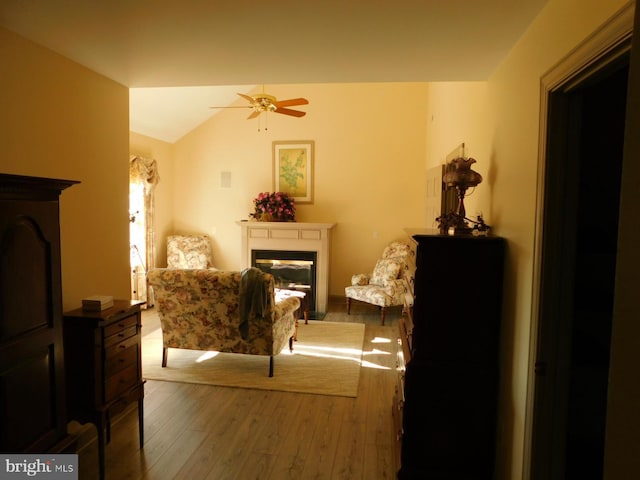 living area with hardwood / wood-style flooring, a glass covered fireplace, vaulted ceiling, and a ceiling fan