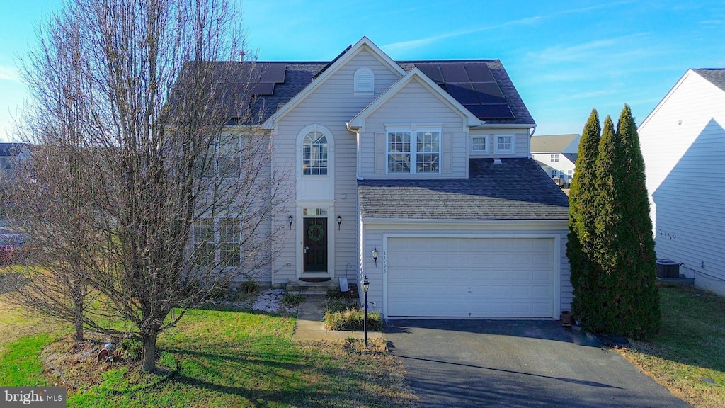 traditional-style house featuring a garage, driveway, a front yard, solar panels, and roof with shingles