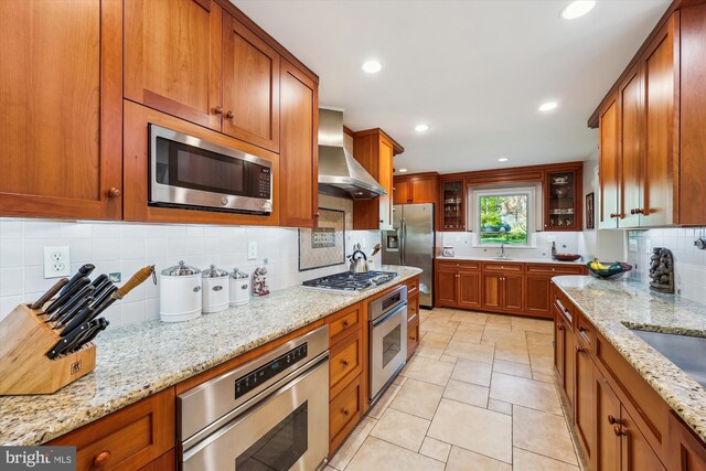 kitchen featuring light stone counters, stainless steel appliances, backsplash, wall chimney range hood, and brown cabinetry