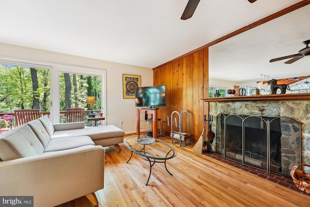 living room featuring a ceiling fan, wood finished floors, a stone fireplace, baseboards, and wood walls