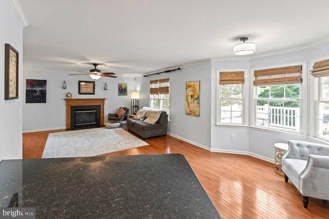 living area with a fireplace, wood-type flooring, crown molding, a ceiling fan, and baseboards