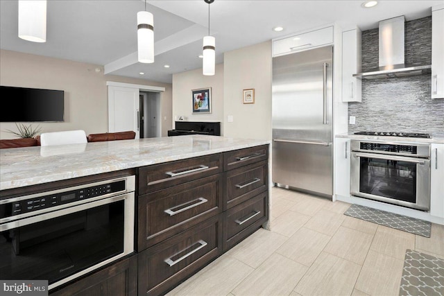 kitchen featuring stainless steel appliances, dark brown cabinets, backsplash, light stone counters, and wall chimney range hood