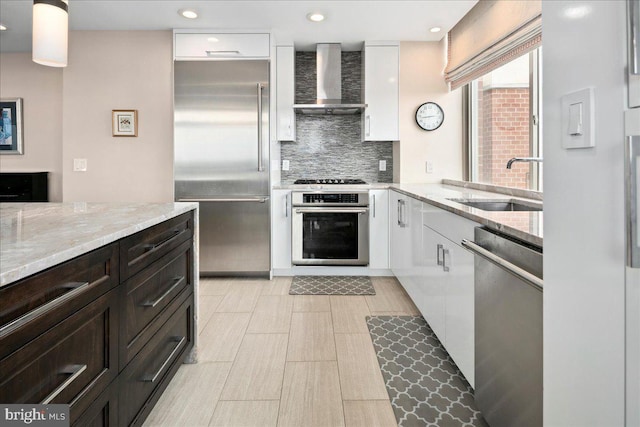 kitchen with stainless steel appliances, backsplash, wall chimney range hood, a sink, and white cabinetry