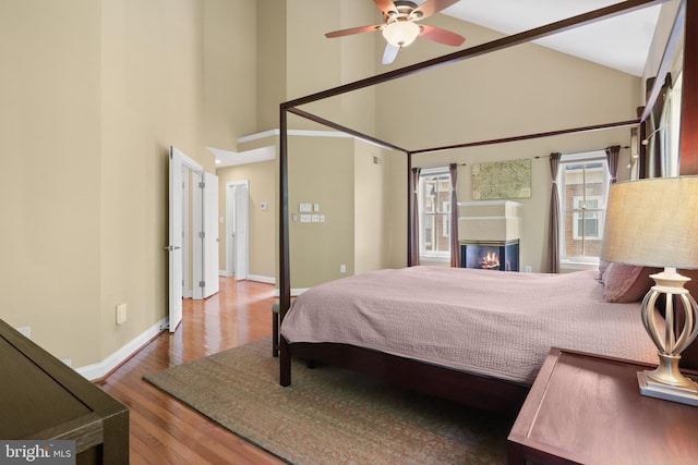 bedroom with baseboards, a glass covered fireplace, wood finished floors, and high vaulted ceiling