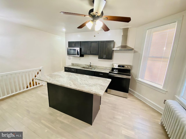 kitchen with radiator heating unit, wall chimney range hood, a ceiling fan, light wood-style floors, and appliances with stainless steel finishes
