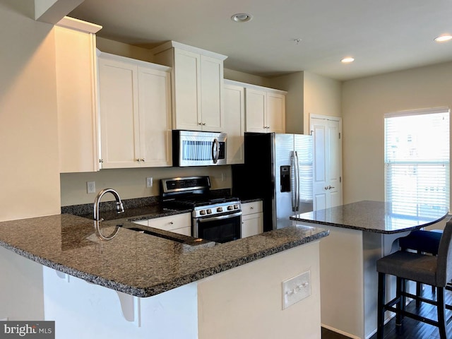 kitchen featuring a kitchen bar, appliances with stainless steel finishes, a peninsula, dark stone counters, and white cabinetry