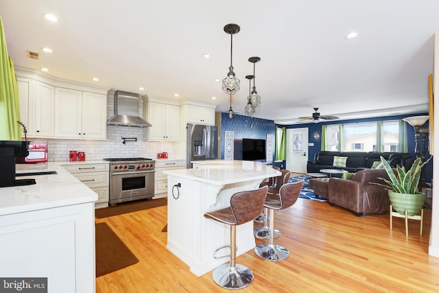 kitchen with wall chimney range hood, ceiling fan, a kitchen island, appliances with stainless steel finishes, and open floor plan