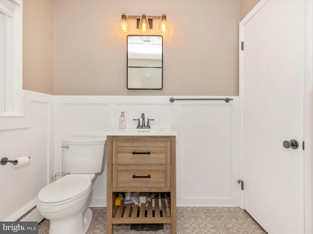 bathroom featuring toilet, tile patterned floors, a decorative wall, vanity, and wainscoting