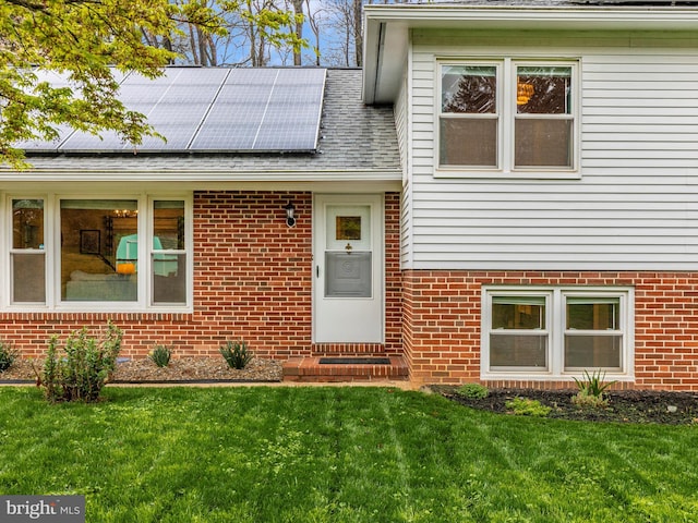 view of exterior entry with a lawn, solar panels, a shingled roof, and brick siding