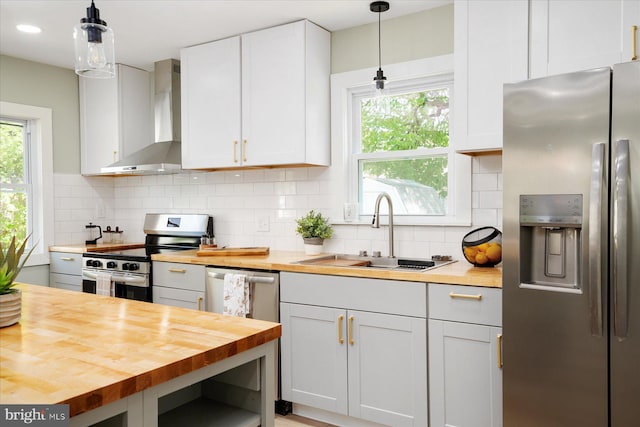 kitchen featuring stainless steel appliances, butcher block countertops, wall chimney exhaust hood, a sink, and pendant lighting