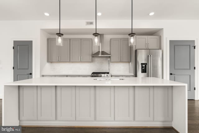 kitchen featuring wall chimney range hood, light countertops, an island with sink, high end refrigerator, and gray cabinetry